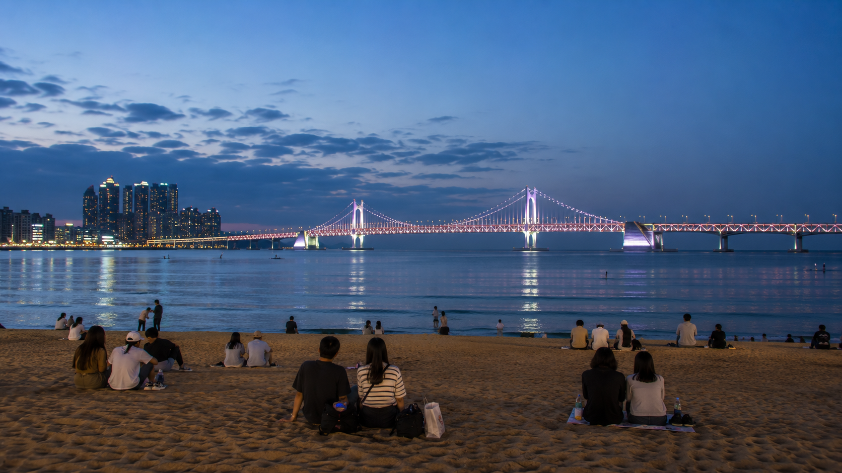 Evening at Gwangalli Beach with people sitting on the sand and Gwangandaegyo Bridge illuminated across the bay